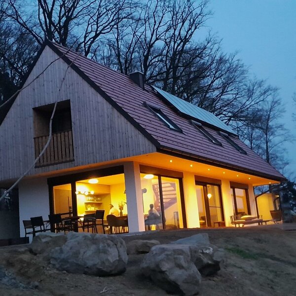 Außenansicht des Bauernhofs in der Abenddämmerung, mit moderner Holzfassade, großen beleuchteten Fenstern im Erdgeschoss, einem Balkon und Solarpaneelen auf dem Dach.