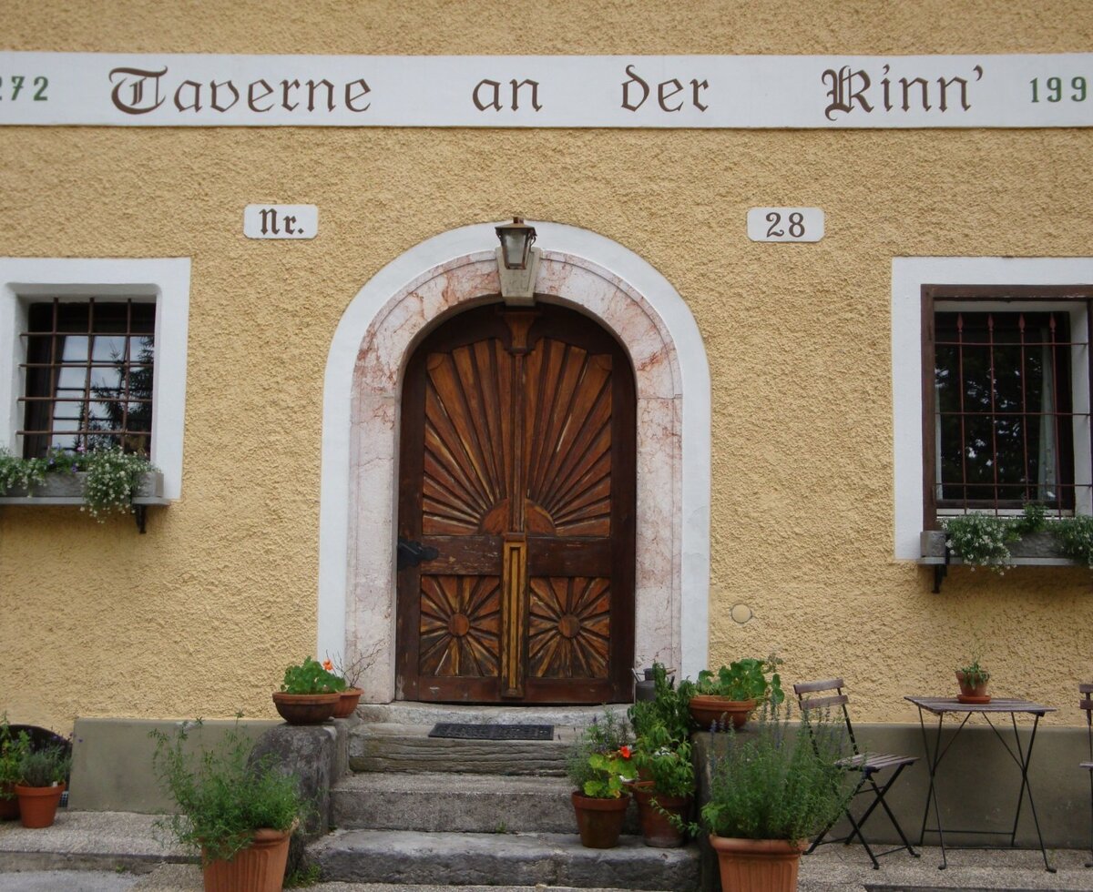 Der Eingang des Bauernhofs mit einer gelben Fassade, einer verzierten Holztür, Fenstern mit Gittern und Blumenkästen sowie einem Schild mit der Aufschrift 'Taverne an der Rinn'.