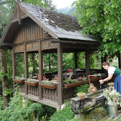 Ein offener Holzpavillon mit Sitzgelegenheiten und ein rustikaler Wasserbrunnen im Garten des Bauernhofs.