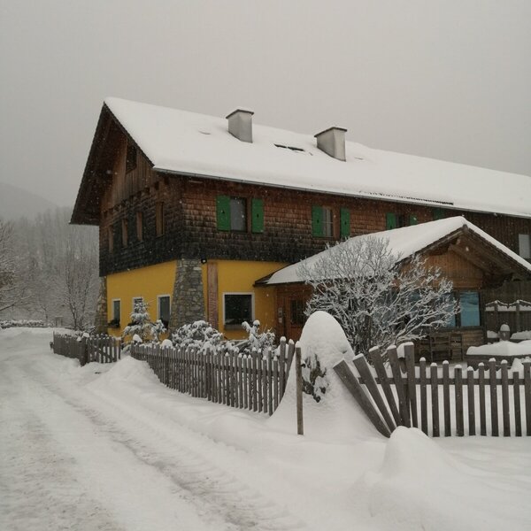 Der Bauernhof im Winter mit schneebedecktem Dach, Holzfassade und einer verschneiten Zufahrt.