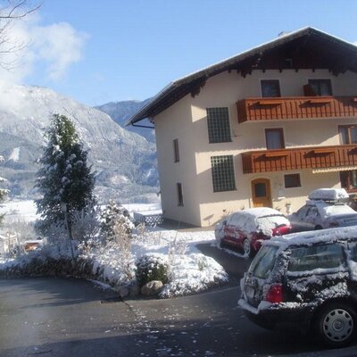 Außenansicht des Bauernhofs im Winter, mit Schnee auf dem Grundstück, parkenden Autos und Bergen im Hintergrund.