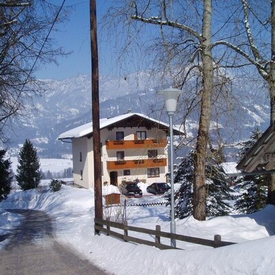 Der Bauernhof im Winter, umgeben von Schnee und Bäumen, mit Blick auf die verschneite Berglandschaft.