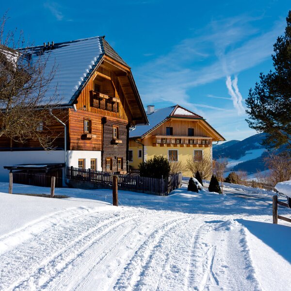 Die winterliche Außenansicht des Bauernhofs mit schneebedeckten Dächern, Zufahrtsweg und der umliegenden Berglandschaft.