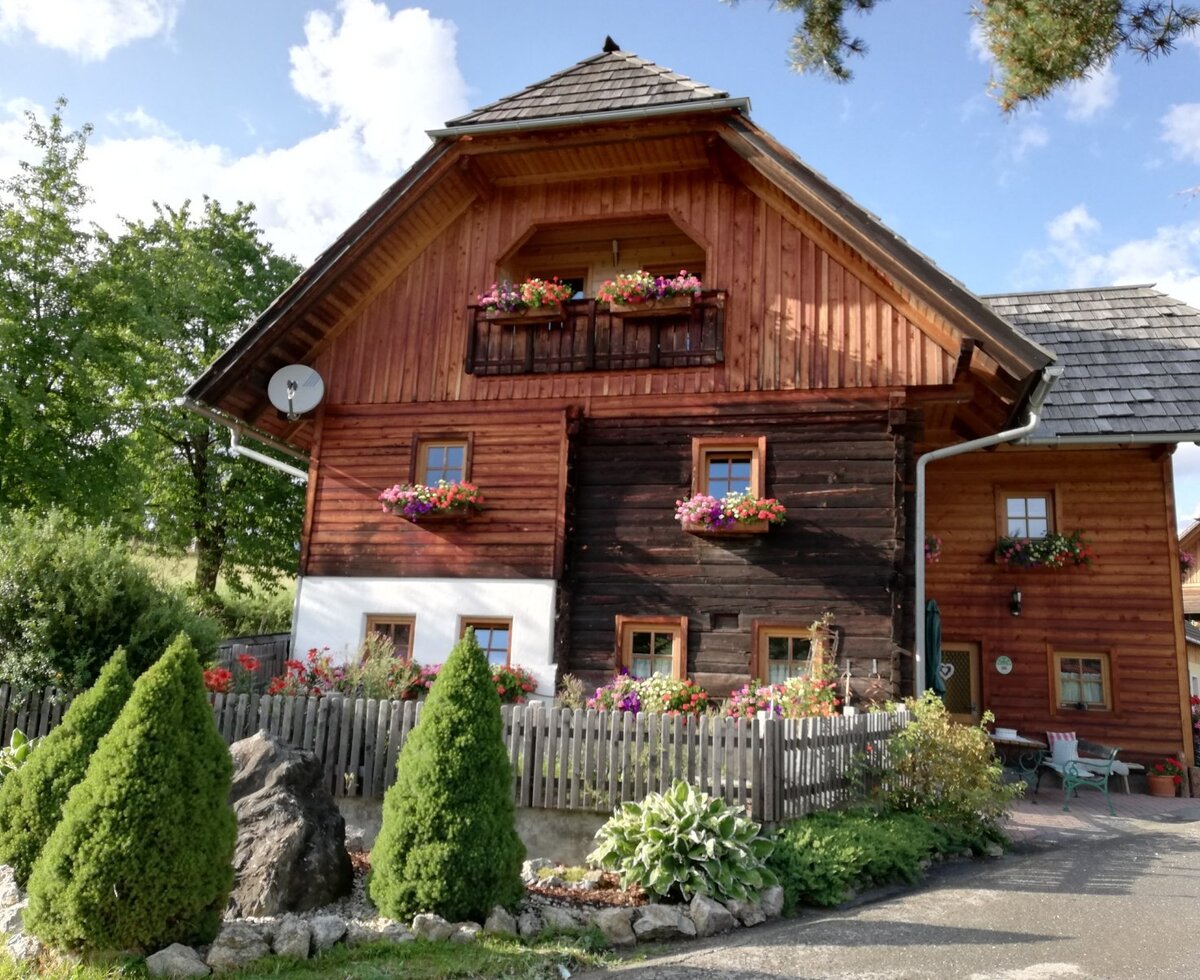 Der Bauernhof von außen mit Holzfassade, Balkon, Blumenkästen an den Fenstern und der Terrasse mit Sitzbereich.
