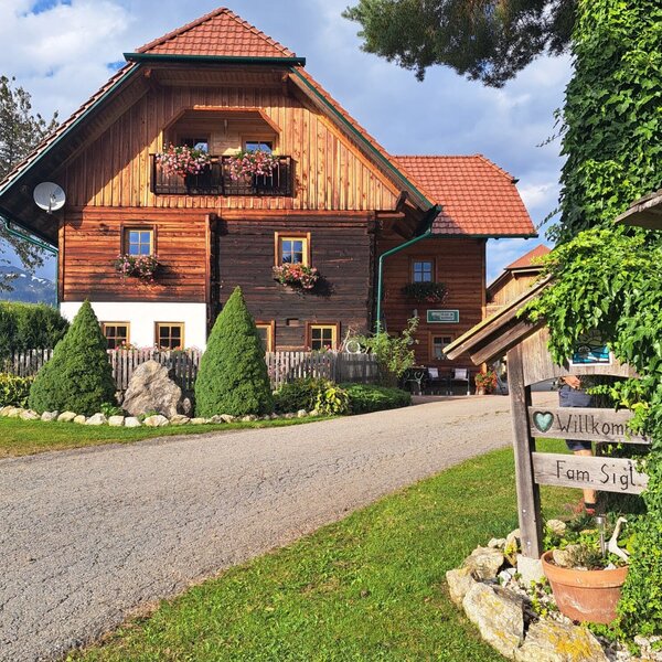 Die Fassade des Bauernhofs mit Holzbalkon, Blumenkästen und einem Willkommensschild am Eingang.