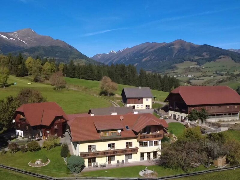 Der Bauernhof mit seinen Gebäuden, umgeben von grünen Wiesen und Wäldern, mit einer Bergkette im Hintergrund.