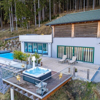 Die einladende Terrasse des Ferienhauses mit einem modernen Whirlpool auf dem Holzdeck, einem Pool und bequemen Liegestühlen vor einer Waldkulisse.