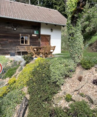 Außenbereich der Hütte mit Holzfassade, weißem Anbau und einer Terrasse mit Holztisch und Bänken.