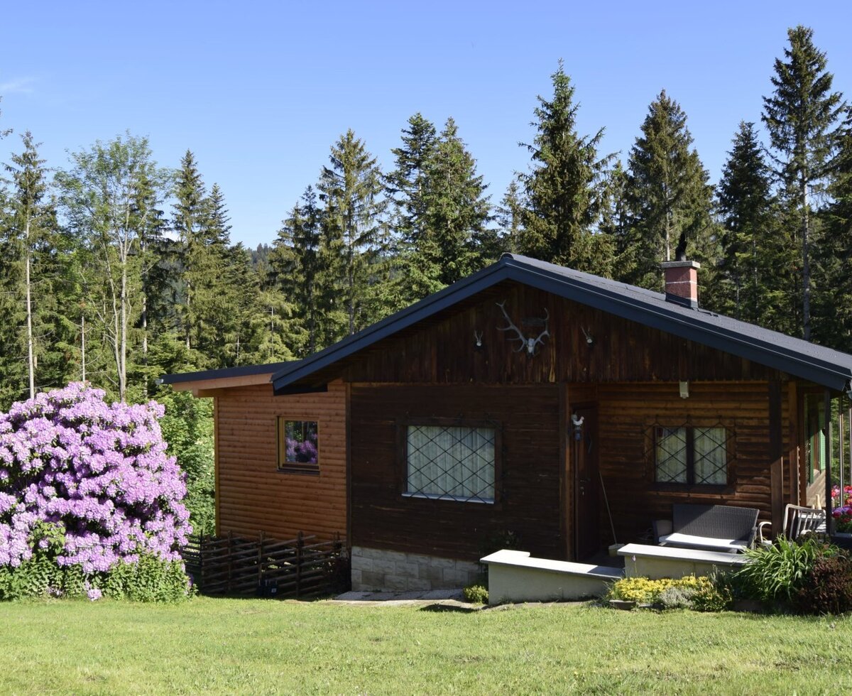 Das Hofgebäude aus Holz mit Gartenfläche und einem blühenden Rhododendronbusch, umgeben von Wald.