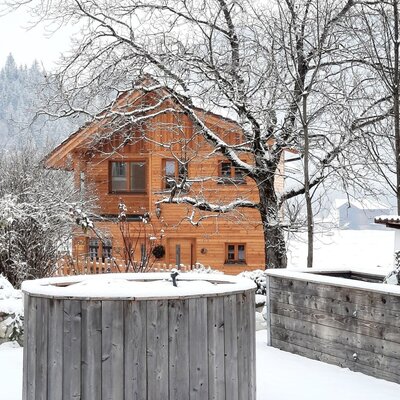 Die Außenseite des Bauernhofs im Winter, mit verschneiten Bäumen und einem hölzernen Badezuber.