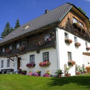 Der Bauernhof mit weiß getünchter Fassade, Holzbalkonen, blumengeschmückten Fenstern und einem Freisitz.