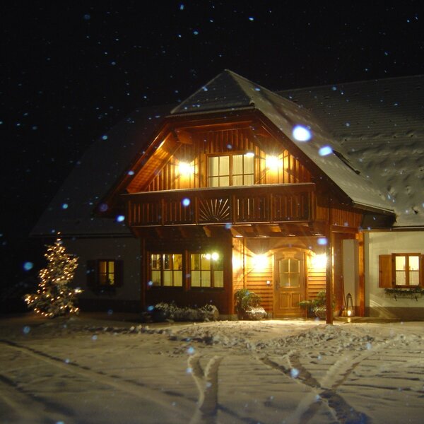 Der beleuchtete Bauernhof bei Nacht, umgeben von Schnee und mit einem geschmückten Weihnachtsbaum.