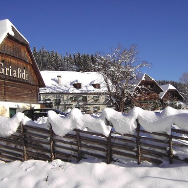 Der winterliche Bauernhof, erkennbar am verschneiten Holzzaun und dem Gebäude mit der Aufschrift "Griaßdi".