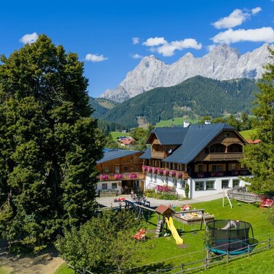 Der Bauernhof mit Kinderspielplatz, Rutsche, Sandkasten und Trampolin, umgeben von grünen Wiesen und Bergen.