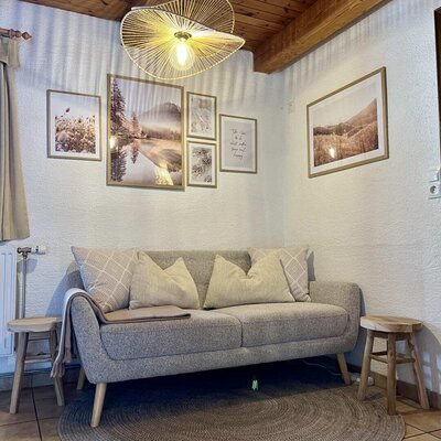 Wohnbereich des Ap­par­te­ments mit grauem Sofa, Kissen, Holzhockern, Wandbildern und einer modernen Deckenleuchte.
