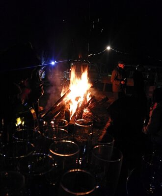 Ein abendliches Lagerfeuer auf dem Bauernhof, umgeben von Gästen und Gläsern.