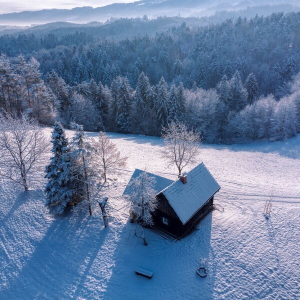 Das Häuschen des Hofes in der verschneiten Winterlandschaft, umgeben von Bäumen und Hügeln.