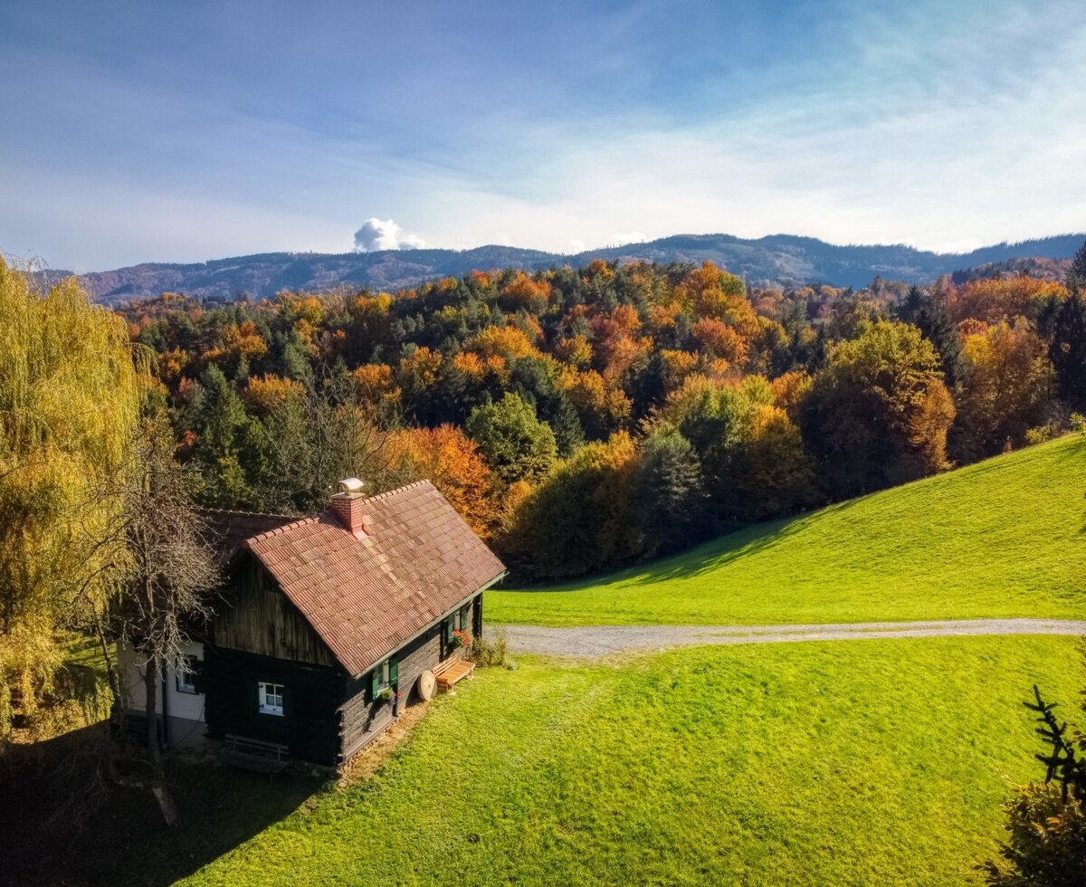 Das Häuschen auf dem Hof liegt auf einem grünen Hügel mit Zufahrtsweg und ist umgeben von herbstlich gefärbten Wäldern.