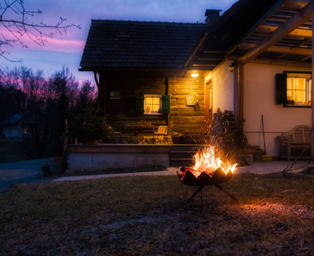 Der Hof mit beleuchteter Fassade, Feuerstelle im Garten und überdachter Pergola am Abend.