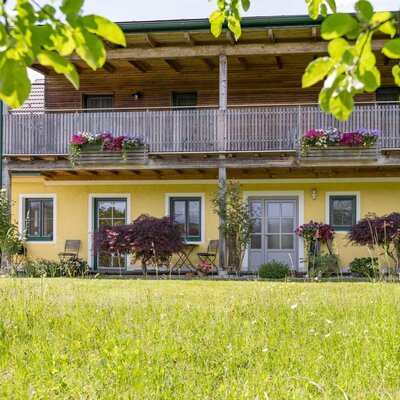 Außenansicht des Bauernhofs mit gelber Fassade, Balkon mit Blumenkästen und ebenerdigem Zugang zu den Doppelzimmern. (c) Elisabeth Fröhlich Photography
