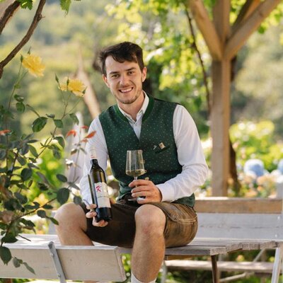 Der Jungwinzer hält eine Weinflasche und ein Glas unter einer Weinlaube im Außenbereich des Hauses.