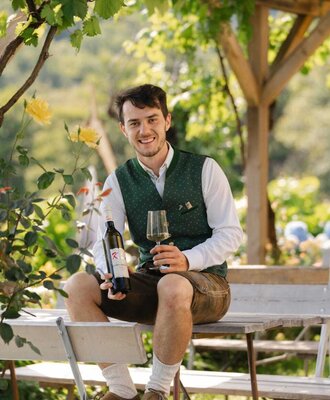 Der Jungwinzer hält eine Weinflasche und ein Glas unter einer Weinlaube im Außenbereich des Hauses.