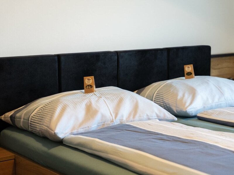 Schlafzimmer mit Doppelbett, dunklem Kopfteil, gestreifter Bettwäsche und kleinen Etiketten auf den Kissen.