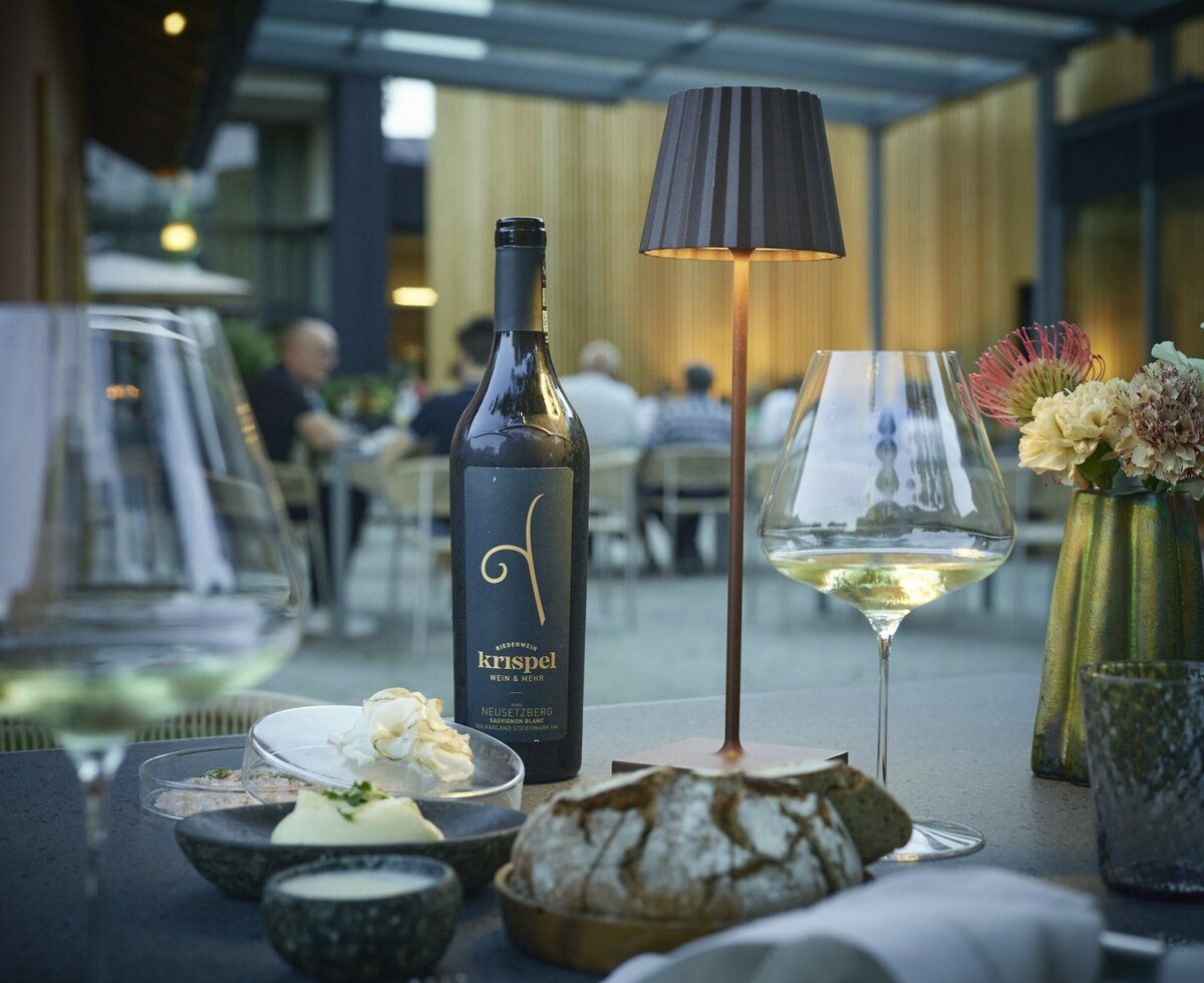 Ein gedeckter Tisch mit einer Flasche Krispel Wein, einem Glas Weißwein und frischem Brot im Gastronomiebereich des Hofes.