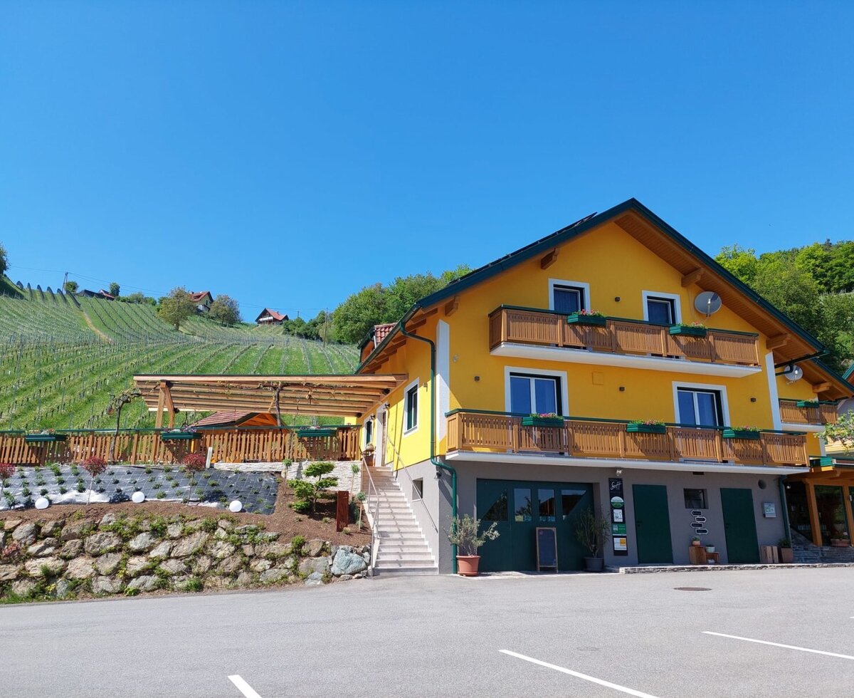 Das Haus mit gelber Fassade, Holzbalkonen und einer Terrasse, gelegen inmitten von Weinbergen, mit Parkplatz.