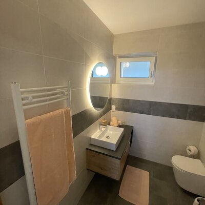 Modernes Badezimmer im Hof mit Waschbecken, beleuchtetem Spiegel, Toilette, beheiztem Handtuchhalter und Fenster.
