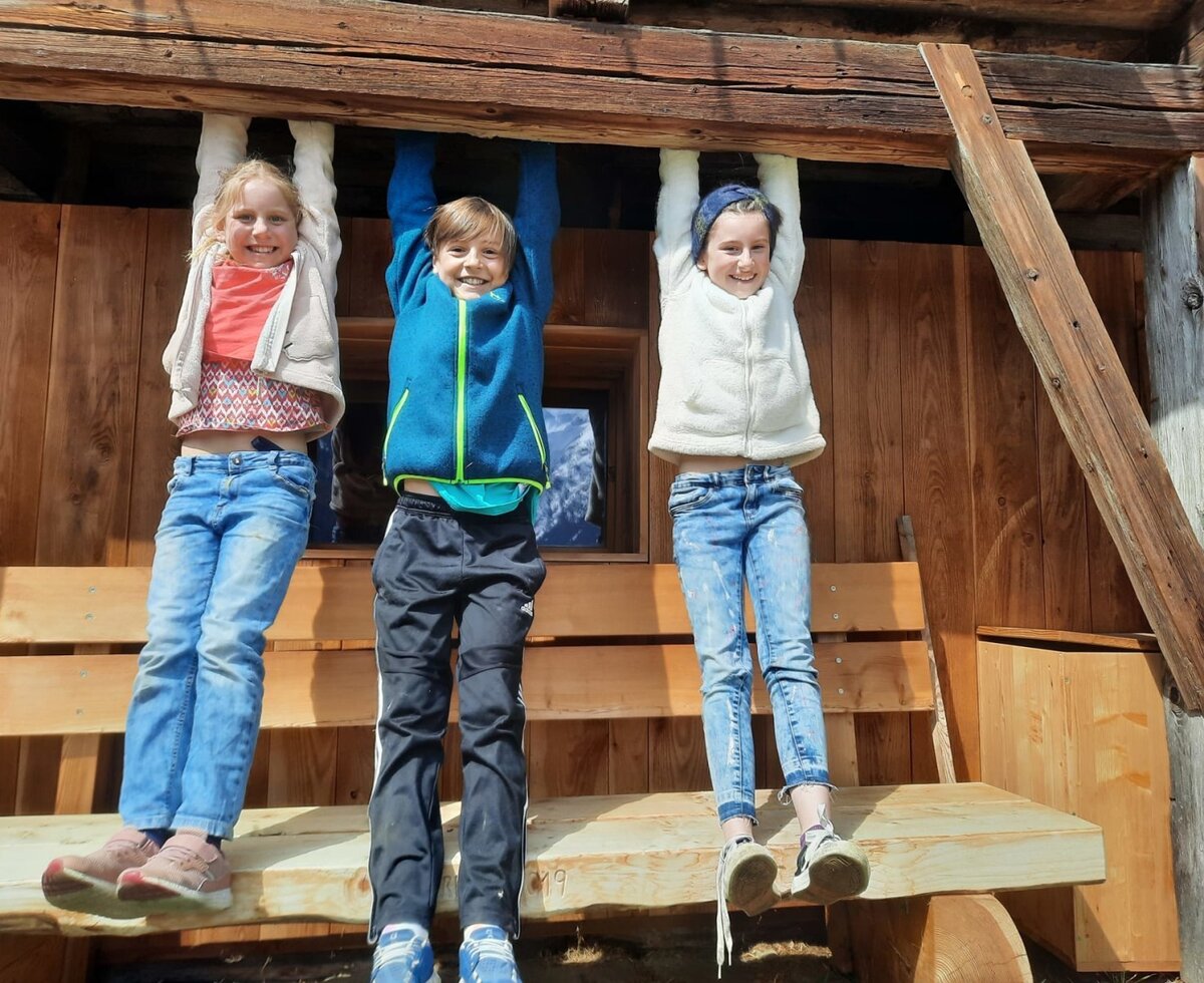 Kinder spielen an einem Holzbalken auf dem Bauernhof, mit einer Holzbank im Außenbereich.