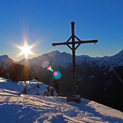 Die winterliche Berglandschaft rund um den Bauernhof, mit einem Gipfelkreuz und strahlendem Sonnenschein.