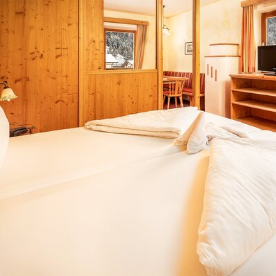 Schlafzimmer einer Suite im Bauernhof mit Doppelbett, SAT-TV, Telefon und einem durch Holzwände abgetrennten Essbereich sowie Fensterblick auf die schneebedeckte Landschaft.