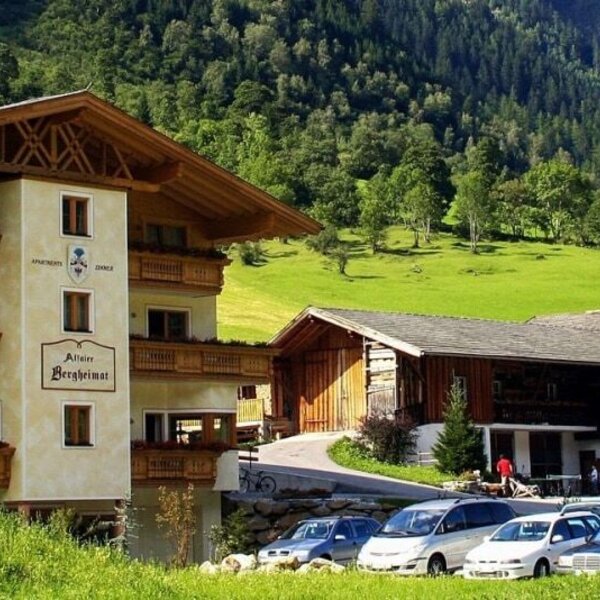 Außenansicht des Bauernhofs mit Balkonen, Parkplatz und Kinderspielplatz vor einer Bergkulisse.