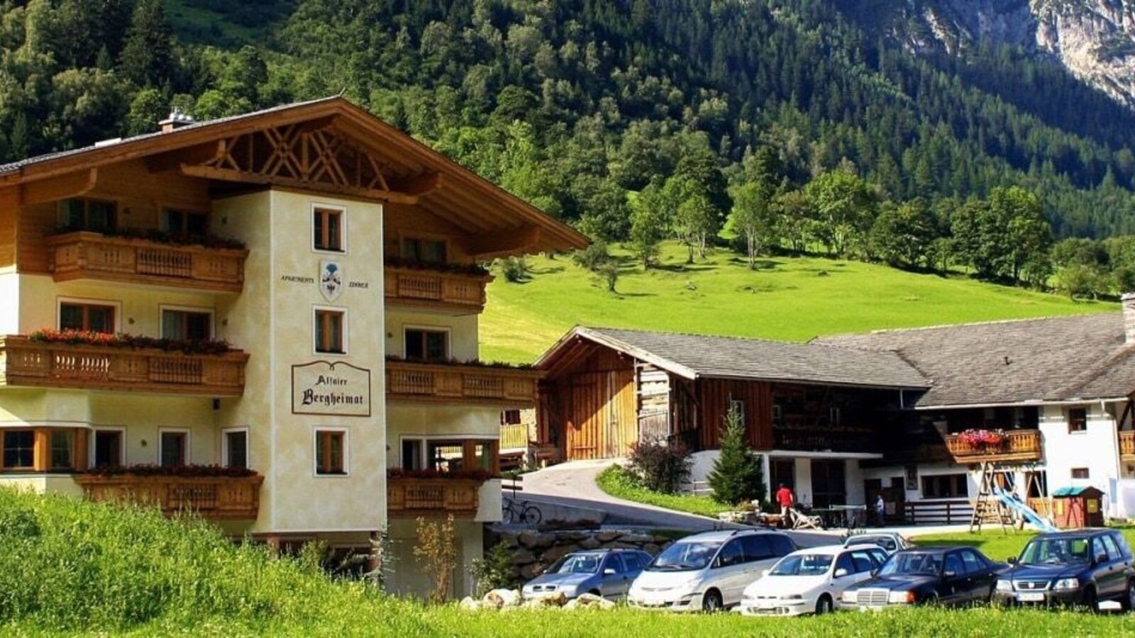 Außenansicht des Bauernhofs mit Balkonen, Parkplatz und Kinderspielplatz vor einer Bergkulisse.