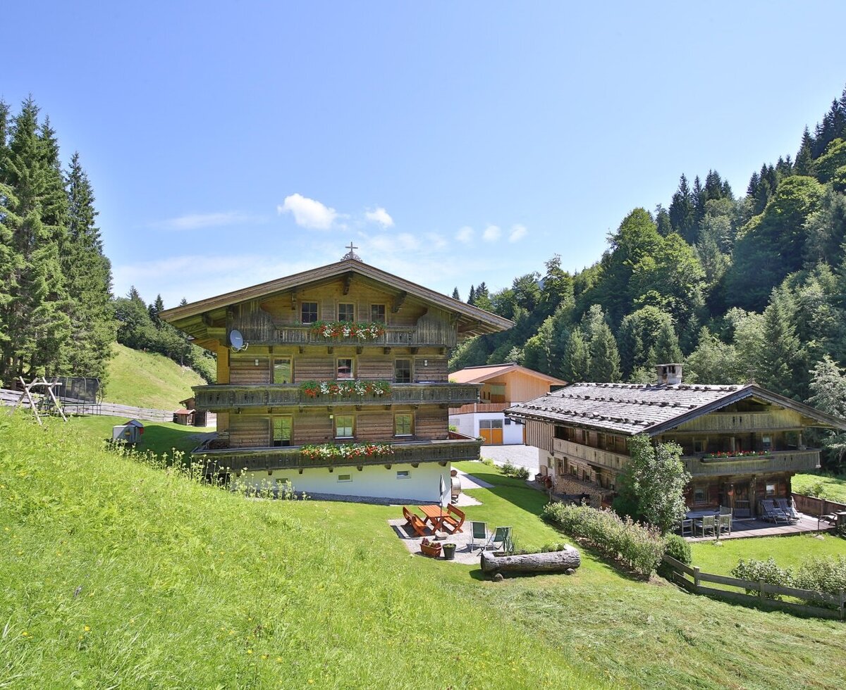 Der Bauernhof als traditionelles Holzhaus mit mehreren Balkonen und einer Terrasse mit Outdoor-Möbeln, umgeben von grünen Hängen und Wäldern.