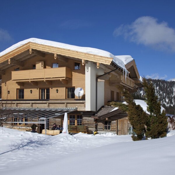 Außenansicht des Bauernhofs im Winter, mit Holzfassade, schneebedecktem Dach, Balkonen und einem überdachten Außenbereich vor Bergkulisse.