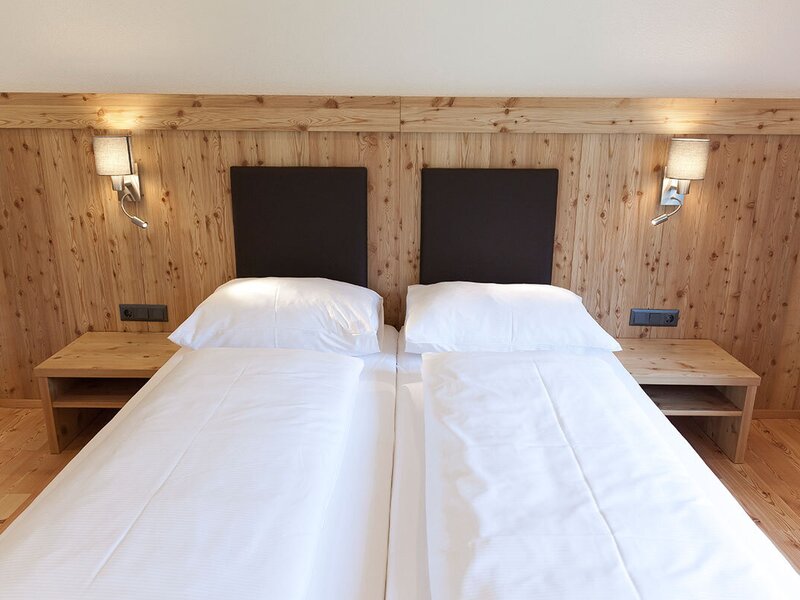 Ein gemütliches Doppelbett im Appartement mit einer rustikalen Holzwand, hellen Nachttischen und einladender Beleuchtung.