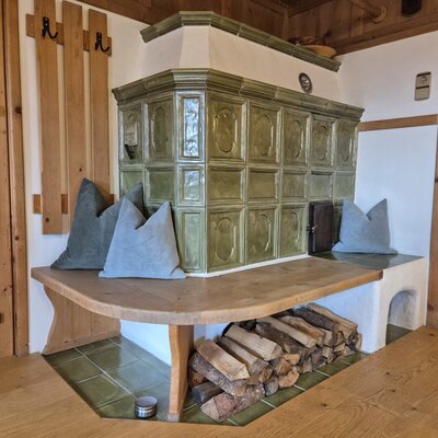 Ein traditioneller, grüner Kachelofen im Bauernhof mit gemütlicher Holzbank, Kissen und ordentlich gestapeltem Brennholz.