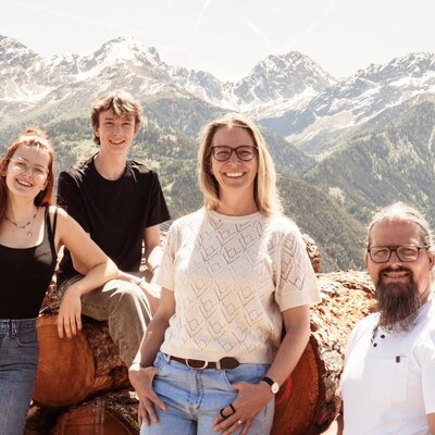 Die Gastgeberfamilie des Bauernhofs vor einer Bergkulisse mit Holzstämmen.