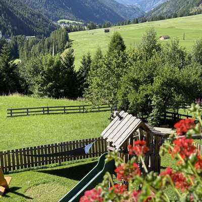 Kinderspielplatz mit Rutsche und Liegestühlen auf dem Rasen des Bauernhofs, mit Blick auf die umliegenden grünen Felder und Berge.