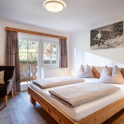 Ein gemütliches Doppelzimmer auf dem Bauernhof mit warmen Holzmöbeln, einem bequemen Sessel und Zugang zum Balkon.