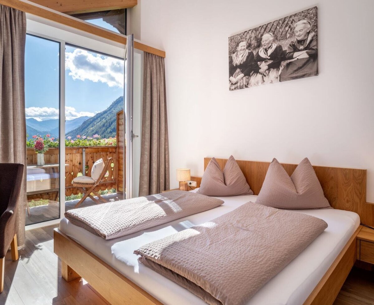Ein gemütliches Doppelzimmer auf dem Bauernhof mit Holzmöbeln, warmen Nachttischlampen und direktem Zugang zum sonnigen Balkon mit Bergblick.