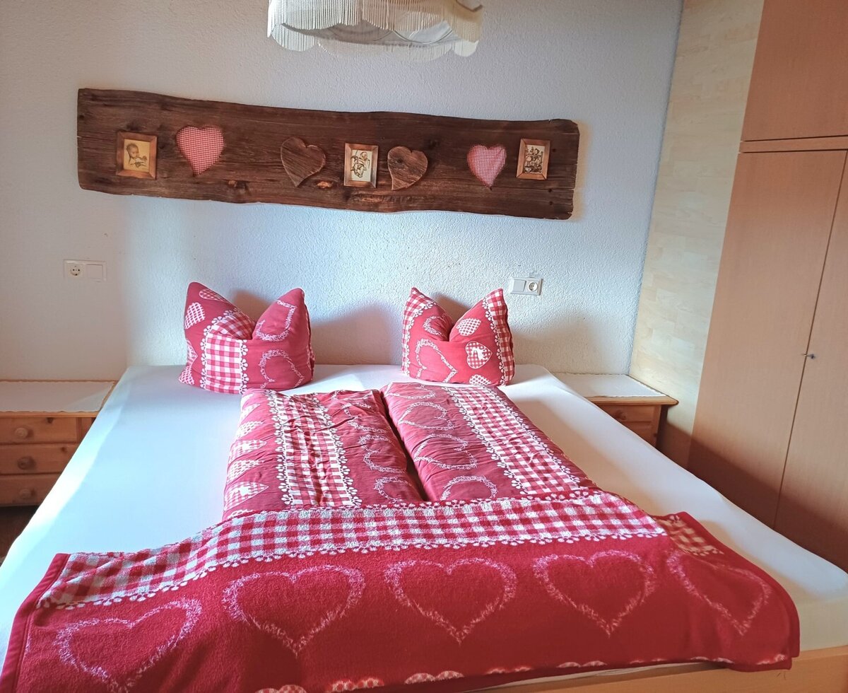 Schlafzimmer in der Alm mit einem Doppelbett, zwei Nachttischen, einem Kleiderschrank und rot-weiß gemusterter Bettwäsche.