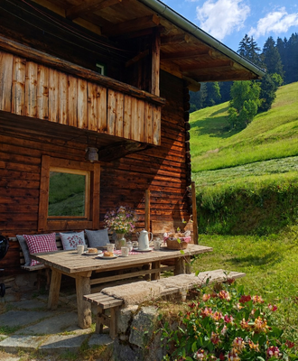 Ein einladender Sitzplatz vor der Alm mit einem rustikalen Holztisch, gemütlichen Kissen und Blick auf die grünen Berghänge.