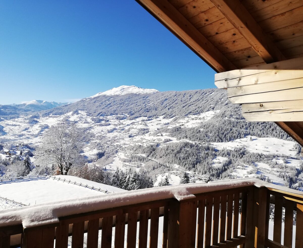 Blick vom Balkon des Bauernhofs auf die winterliche Berglandschaft mit verschneiten Tälern und einem klaren blauen Himmel.