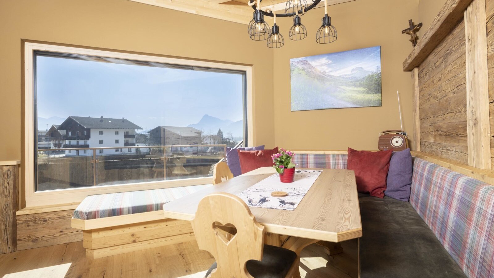 Essbereich im Bauernhof mit Eckbank, Holztisch und großem Fenster mit Aussicht auf die umliegenden Häuser und Berge.