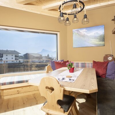 Essbereich im Bauernhof mit Eckbank, Holztisch und großem Fenster mit Aussicht auf die umliegenden Häuser und Berge.
