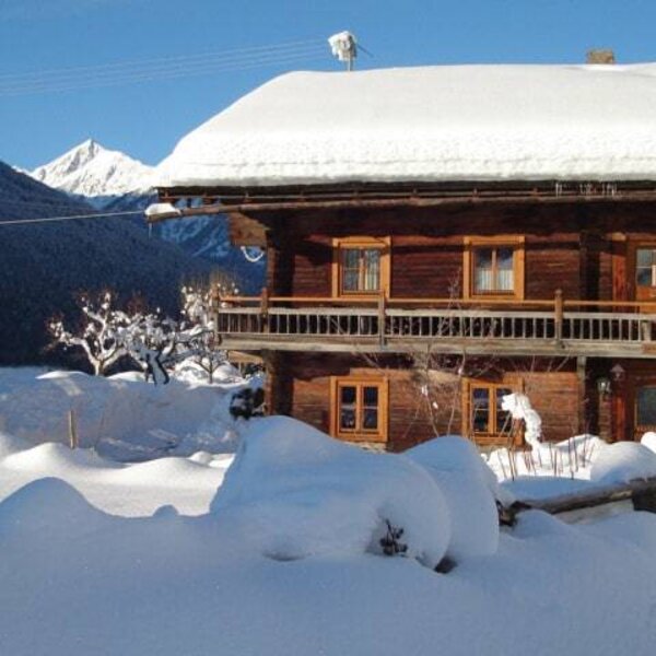 Der Bauernhof mit schneebedecktem Dach, Holzbalkon und winterlicher Berglandschaft.