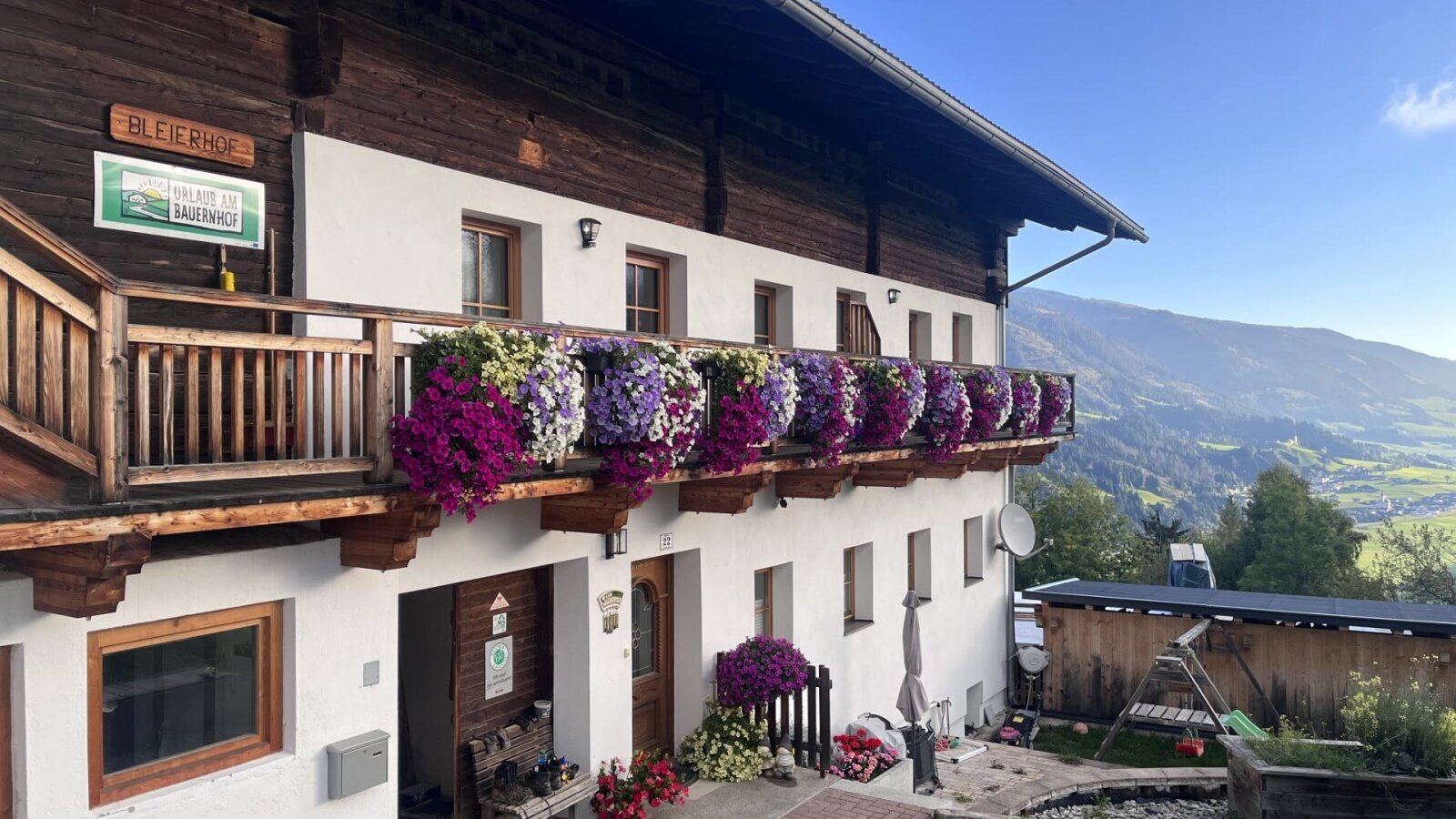 Außenansicht des Bauernhofs mit blumengeschmücktem Balkon, Bergblick und Kinderschaukel im Garten.